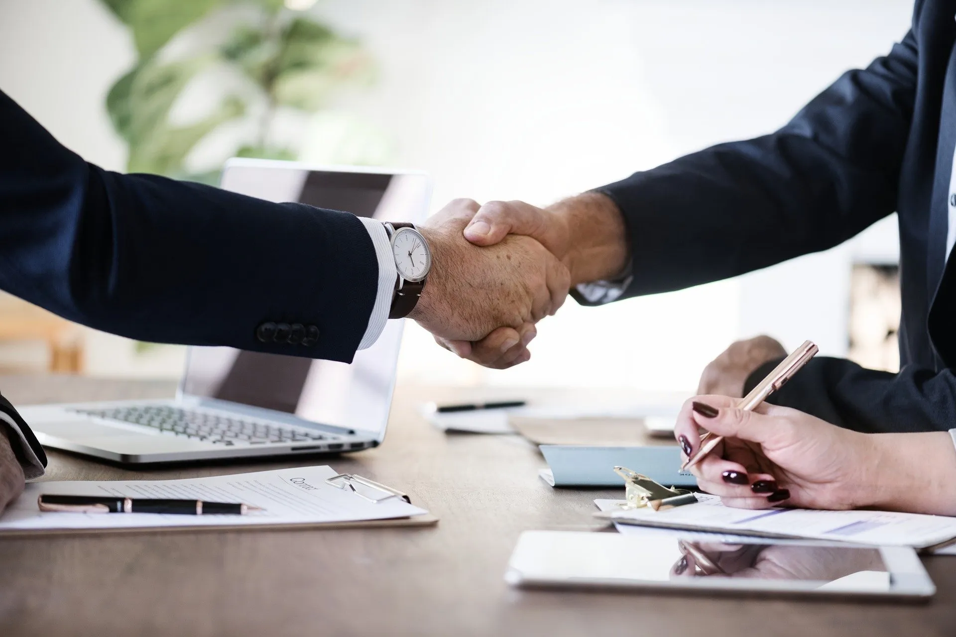 Two business professionals shaking hands over a desk with a laptop and paperwork